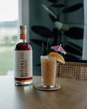 A bottle of RITUAL ZERO PROOF- RUM ALTERNATIVE by RITUAL ZERO PROOF, featuring a tan cap, amber liquid, and a white label with bold black and orange text describing this non-alcoholic rum alternative.