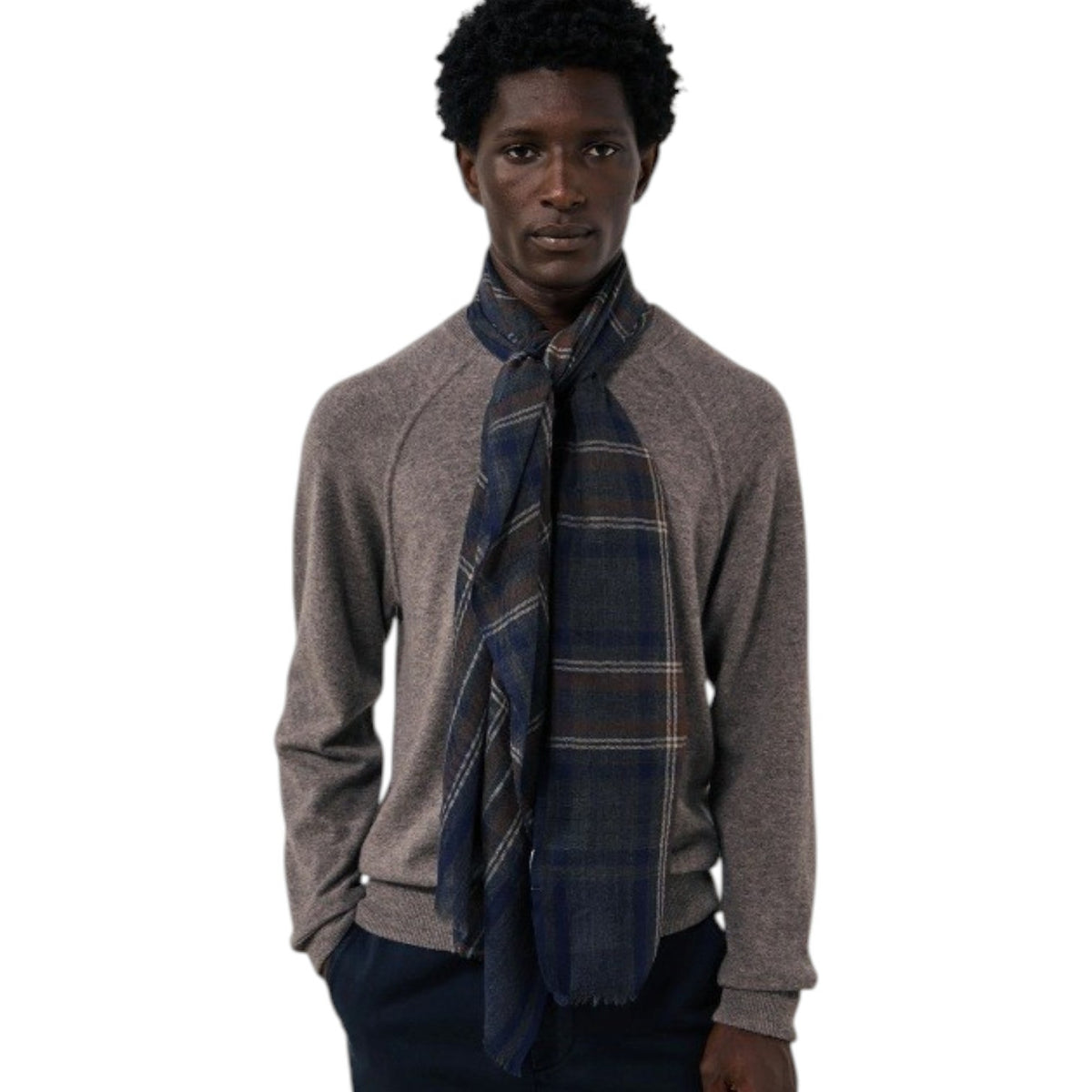 A man wearing a grey long-sleeve sweater and the HARTFORD - TARTAN WOOL SCARF IN INDIGO, GREY AND CAMEL stands against a white background, looking directly at the camera with his hands in his pockets—showcasing this classic men’s winter accessory from HARTFORD.