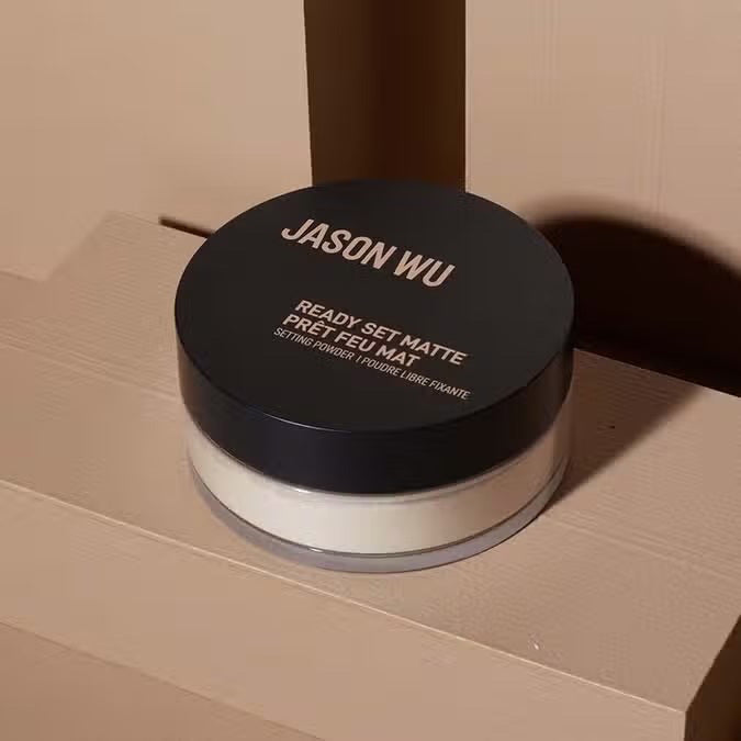 A container of JASON WU BEAUTY - READY SET MATTE 1 - TRANSLUCENT BANANA setting powder sits on a light brown textured surface. The cylindrical container has a black lid with the brand name in white capital letters and product details in both English and French, ensuring a flawless matte finish for any makeup look.