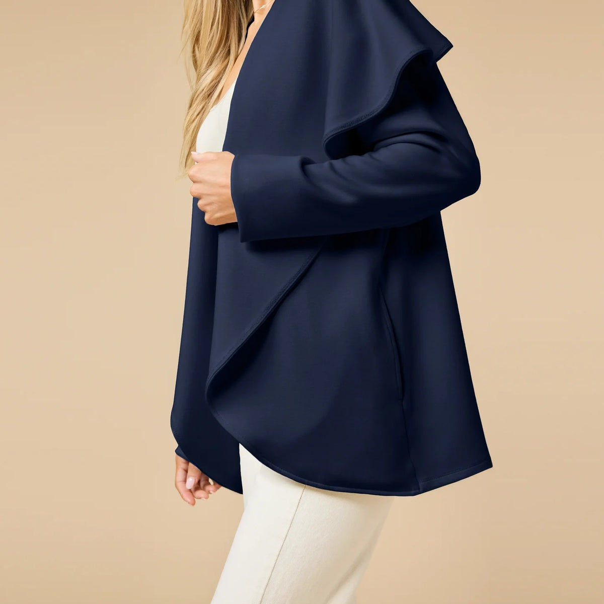 A woman wears an off-white SOFTIES - DREAMTECH EXAGGERATED COLLAR CARDIGAN with an exaggerated waterfall collar, cinched at the waist with a thin belt, and holds a matching handbag.