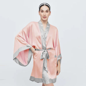 The BRANCHE GEISHA Robe Silk in pink satin, adorned with white trim, hangs gracefully on a matching silk hanger. This elegant robe by BRANCHE features long, wide sleeves and is accentuated with a white sash tied in a bow around the waist, complete with mitered corners for an exquisite finish.