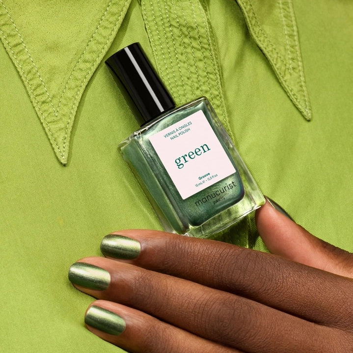 A hand with metallic green nails holds a bottle of MANUCURIST - GREEN™ NAIL POLISH IN GROOVE from MANUCURIST INC, perfectly matching the light green shirt in the background.