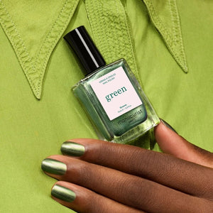 A hand with metallic green nails holds a bottle of MANUCURIST - GREEN™ NAIL POLISH IN GROOVE from MANUCURIST INC, perfectly matching the light green shirt in the background.