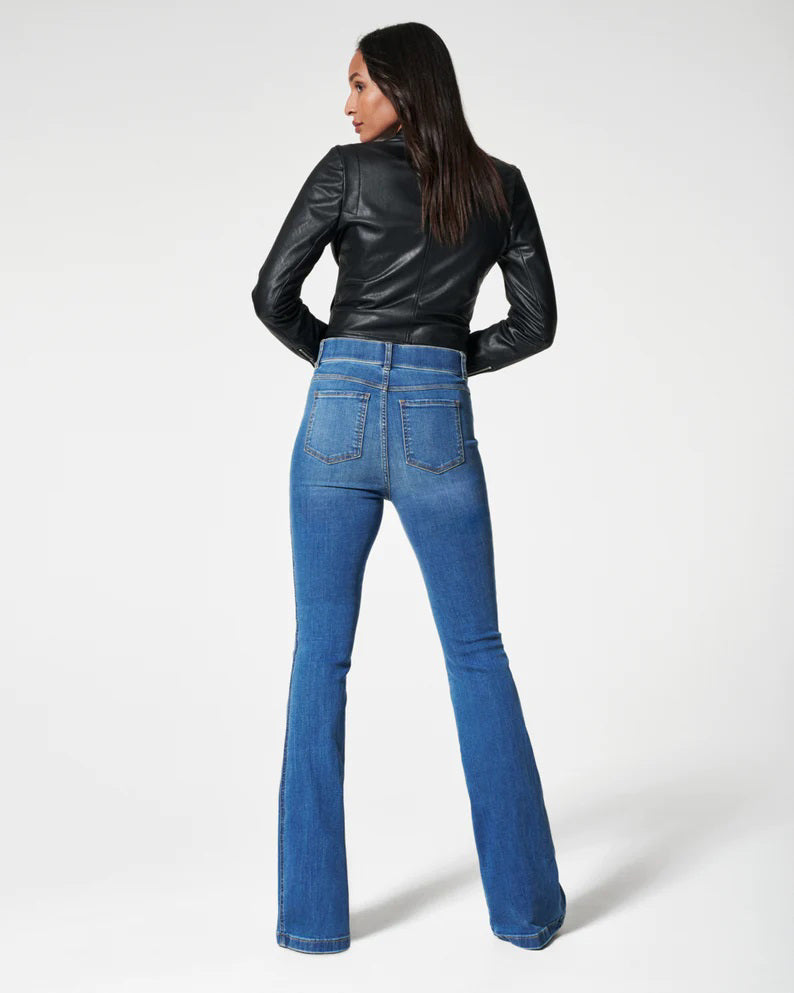 A woman poses against a white background in the SPANX jeannish high-waisted blue flare jeans made from premium stretch denim with hidden core shaping technology, styled with a black faux leather jacket, black top, and pointed-toe shoes.