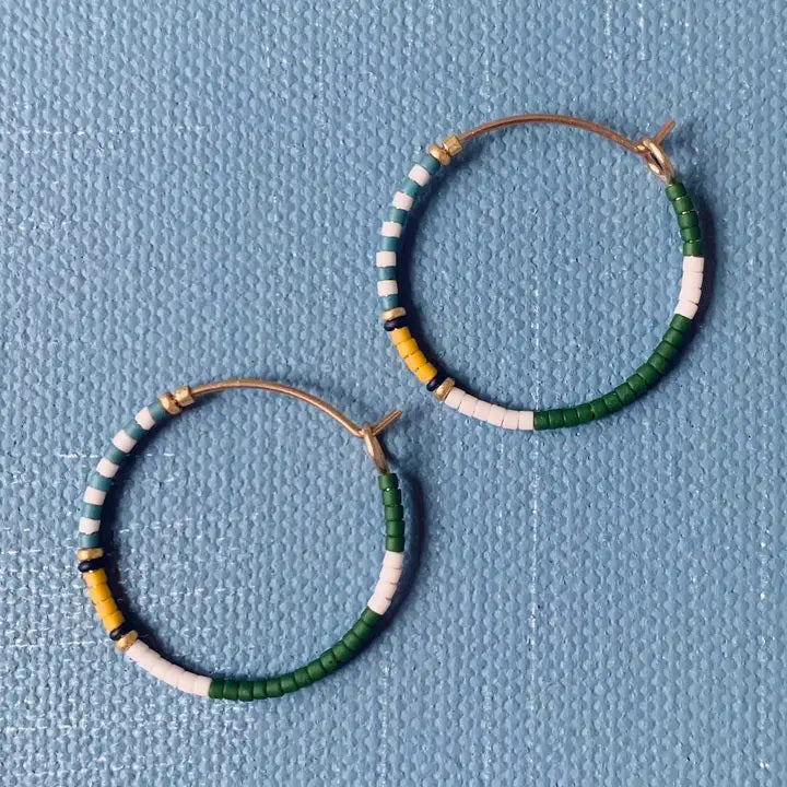 The ALICE RISE MIDI COLORLOOP EARRINGS IN FAIRWAY GREEN feature round 14k gold filled hoops with green, white, yellow, and black Japanese glass beads, displayed on a textured light blue background.