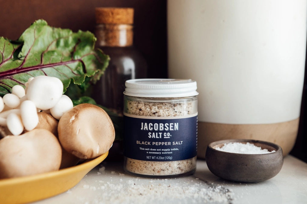 A small glass jar of JACOBSEN SALT CO. JACOBSEN SALT - INFUSED BLACK PEPPER SALT with a white lid rests on a light beige surface against a light gray background. The dark label with white text highlights this floral, spicy blend.