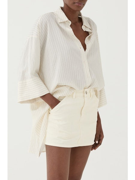 A person wears the ELEH STRIPED SHIRT WITH COLLAR, an oversized, cream pinstripe top with wide sleeves, partially tucked into a high-waisted cream mini skirt, standing against a plain white background.