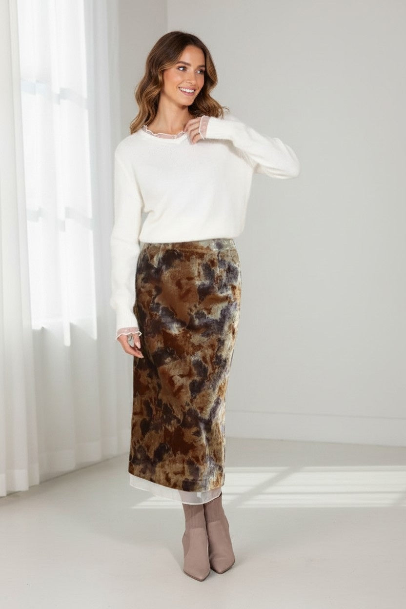 A woman stands in a bright room, sunlight streaming through white curtains, smiling and touching her hair. She wears a white sweater paired with the JESSIE LIU Printed Velvet Midi Skirt with Organza Trim—a floor-grazing abstract print skirt from JESSIE LIU—and beige ankle boots.