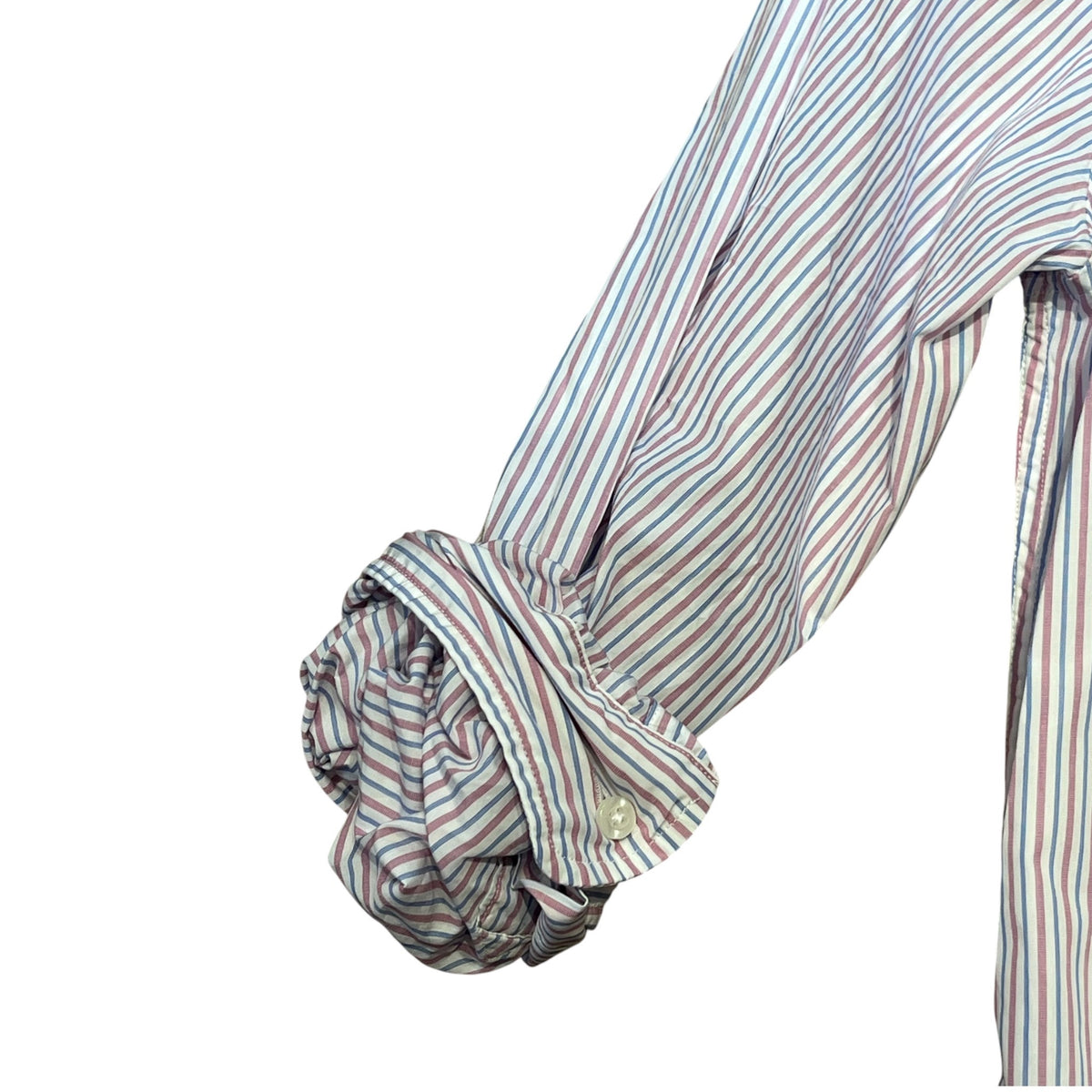 A man with short gray hair and a beard sits on a park bench wearing the FRANK & EILEEN Finbar men's button-up shirt in pink and blue multi stripe, blue jeans, and white sneakers. Behind him, people walk along a flower-lined path among trees.