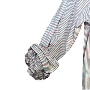 A man with short gray hair and a beard sits on a park bench wearing the FRANK & EILEEN Finbar men's button-up shirt in pink and blue multi stripe, blue jeans, and white sneakers. Behind him, people walk along a flower-lined path among trees.