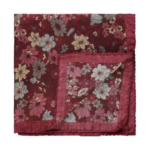 The AMANDA CHRISTENSEN Silk Pocket Square with Floral Pattern in Wine Red Melange is shown folded, highlighting its maroon silk fabric adorned with pink, yellow, and gray floral motifs. One corner is turned to reveal the reverse side, illustrating its quality craftsmanship and making it a sophisticated addition to any men’s accessory collection.