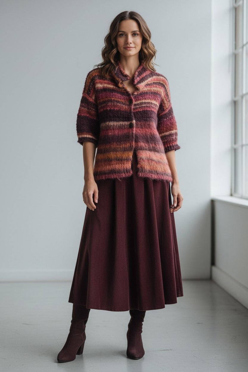 A woman with wavy hair and a calm expression stands indoors by a window, dressed in the FLOOR Corduroy Skirt in Pomegranate, a striped multicolored knit cardigan, and matching boots. The setting is minimal and bright.