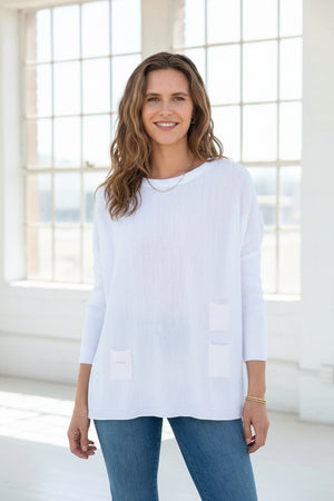 A smiling woman with curly hair wears the MERSEA - CATALINA CREWNECK SWEATER in navy blue and jeans, standing against a plain light background with her hands near her head.