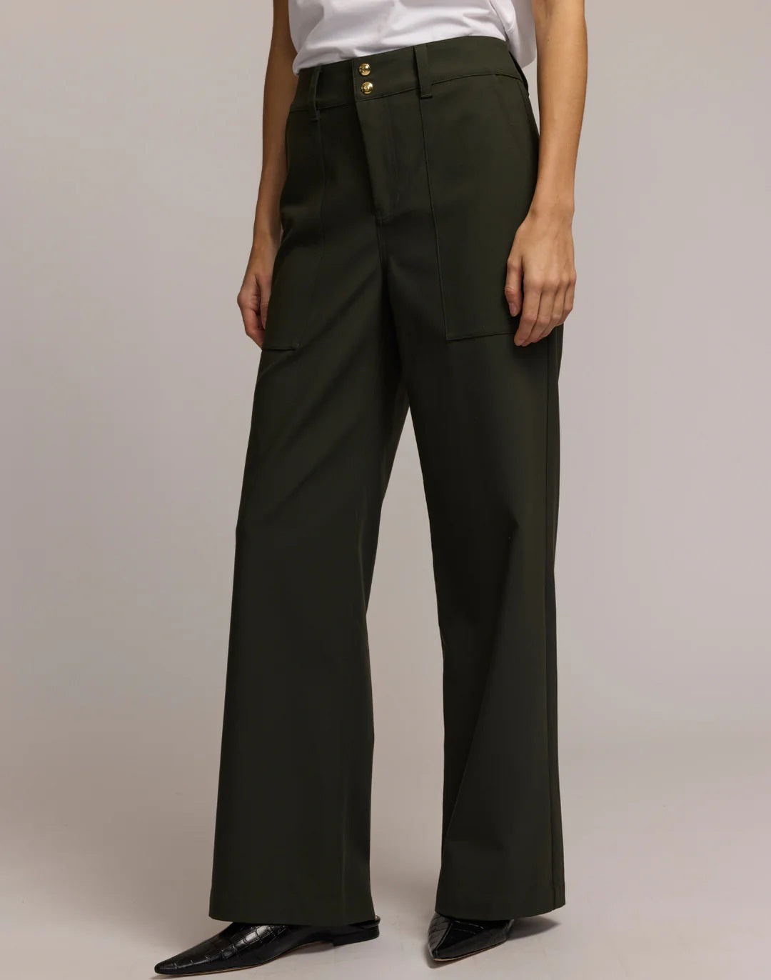 A person wears HINSON WU&#39;s KELLY TRAVELER&#39;S TWILL PANT in dark green with front pockets and gold buttons, styled with a white top and black pointed-toe shoes. Photo is cropped from shoulders down, highlighting this travel-ready look.