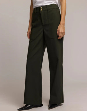 A person wears HINSON WU's KELLY TRAVELER'S TWILL PANT in dark green with front pockets and gold buttons, styled with a white top and black pointed-toe shoes. Photo is cropped from shoulders down, highlighting this travel-ready look.