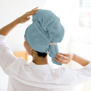 A white cylindrical VOLO BEAUTY - HERO HAIR TOWEL LUXE container stands beside a rolled, absorbent blue towel with a light gray tag and strap. Both feature the VOLO BEAUTY logo and are designed for frizz-free drying.