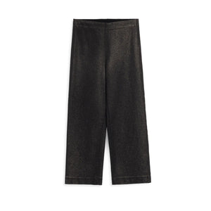 A woman stands in a doorway wearing a light blue shirt and FRANK & EILEEN’s WESTPORT ILLUSION Wide Leg Pull-On Pant in Black Gold Glitter. A cozy living room with a sofa, lamp, and plants is visible in the background.