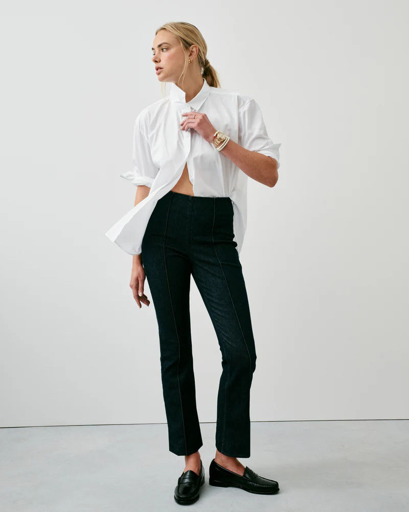 A woman stands against a white background wearing SPANX&#39;s ZERO WAISTBAND MICRO FLARE JEAN in Raw Indigo Wash, paired with a white top, tan heeled sandals, a gold bracelet, and a gold ring.