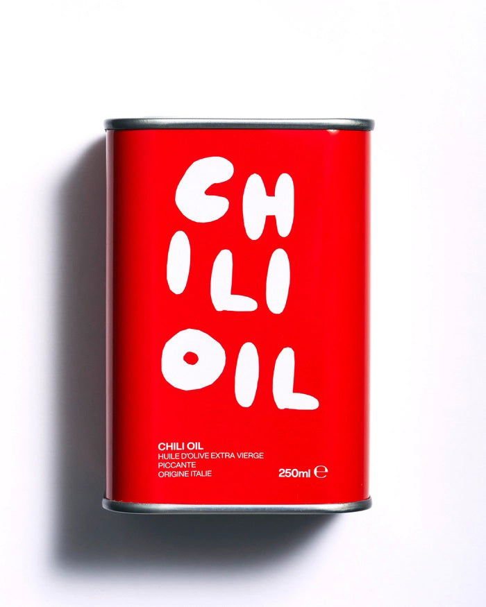A red rectangular can with a black top and bottom, featuring the product name &quot;OLEA PIA - CHILI OIL&quot; in large white letters at the top. Below, in smaller white text, it reads: &quot;Super Spicy Oil, Huile d&#39;Olive Extra Vierge, Piccante, Origine Italie, 250ml,&quot; from the brand OLEA PIA.