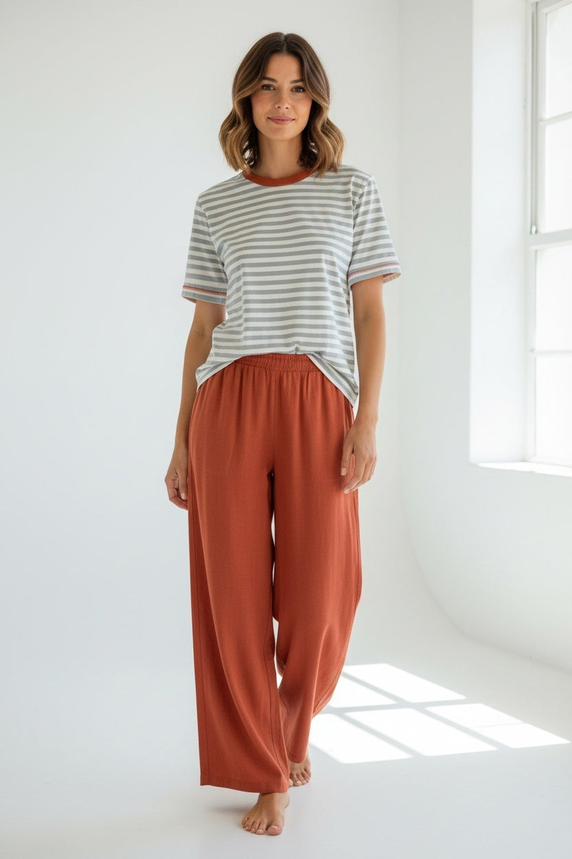 Wearing the MERSEA Deauville Ringer Tee in Slate Blue Persimmon and red pants, a woman sits on white stone steps beside a blue door, holding a brown MERSEA bag.