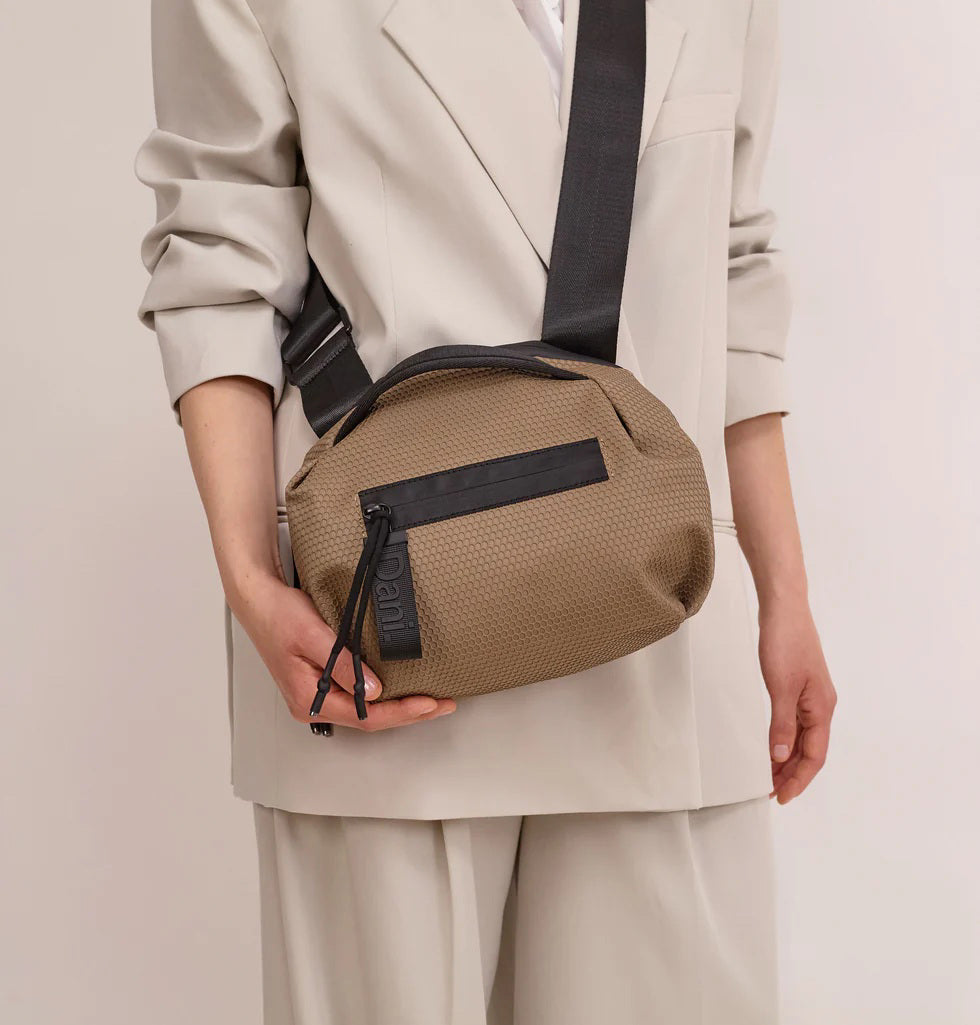 A person in a light beige suit carries the DANIELLA LEHAVI - BONNIE TRAVEL CROSSBODY, a large brown vegan bag with black zipper and strap, shown from shoulders to mid-thigh against a plain background.