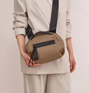 A person in a light beige suit carries the DANIELLA LEHAVI - BONNIE TRAVEL CROSSBODY, a large brown vegan bag with black zipper and strap, shown from shoulders to mid-thigh against a plain background.