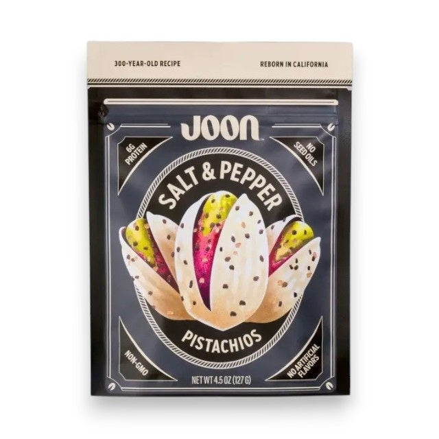 A bag of JOON- SALT & PEPPER PISTACHIOS by JOON, a savory plant-based snack, rests on a white tiled surface with pistachios and a bowl of salt nearby. The packaging features claims such as non-GMO, no artificial flavors, and 6g of protein per serving.