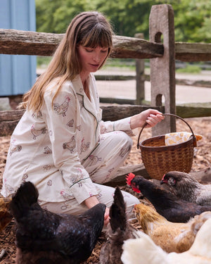 A woman dressed in the PRINTFRESH Embroidered Hens Long Pajama Set in Buff smiles as she stands by a wooden fence, holding a basket and watching chickens peck at the ground in a farmyard setting, with trees and a path visible behind her.