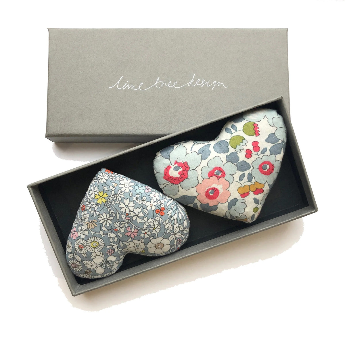 A grey rectangular box labeled with &quot;LIME TREE COLLECTION LTD&quot; on the lid contains two heart-shaped sachets from the HEARTS - LOVE HEART SACHET collection. Crafted from Liberty Tana Lawn fabric, one sachet features a blue floral pattern while the other has a white background adorned with colorful flowers. Both are filled with fragrant lavender.