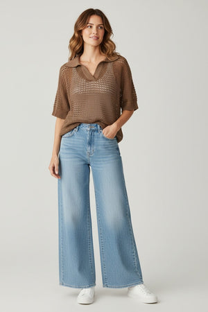 A woman stands against a plain background, smiling slightly with one hand in her pocket. She wears the BASE MILANO - SHORT SLEEVE SPLITNECK COTTON POLO IN BROWN over a tan top, paired with light blue wide-leg jeans and white sneakers. Her wavy brown hair frames her face as the breathable texture of the BASE MILANO sweater lends an effortless touch to her relaxed look.