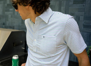 A man with dark curly hair smiles in the CRIQUET Top-Shelf Players Shirt in Stewart Stripe White, featuring light blue and white stripes, a chest pocket, and dark jeans, standing against a white background with one hand in his pocket.