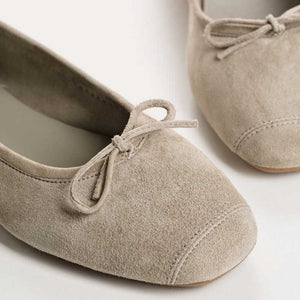 The REQINS - HARMONY BALLERINA FLATS by REQINS are shown in taupe suede with a small front bow and low heel, displayed in side profile against a plain light background.