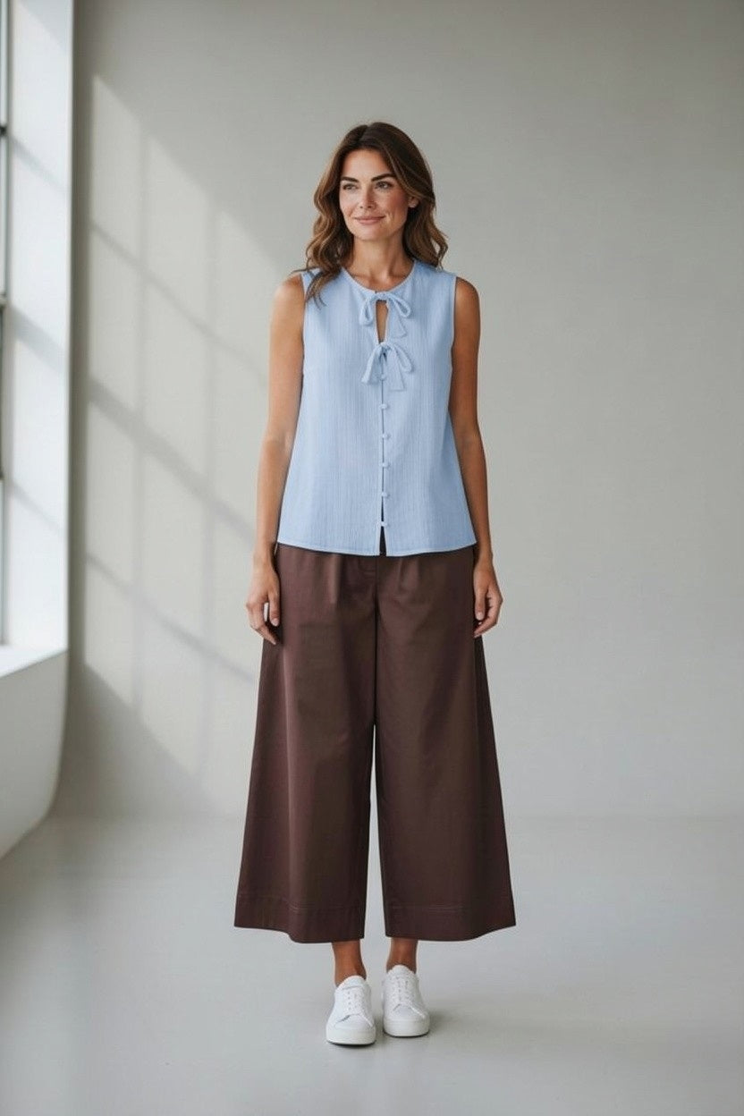 A woman with wavy hair smiles softly indoors, wearing the SANDRO FERRONE Tie Front Sleeveless Top in Celeste paired with brown wide-leg pants and white sneakers. The plain background features soft natural light.