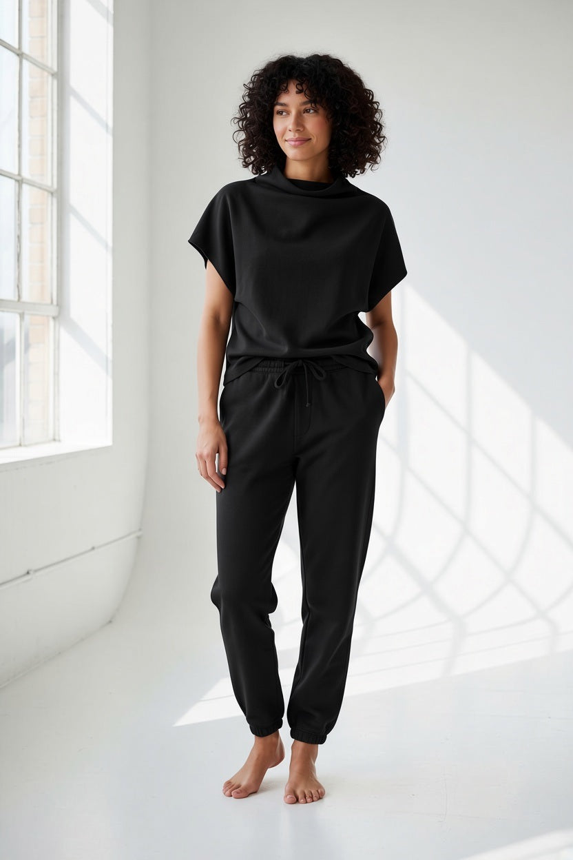 A woman with blonde hair in a ponytail wears the MERSEA Frenchie Dolman Top in Black and black joggers, sitting casually on a silver suitcase against a plain beige wall, effortlessly showcasing the relaxed silhouette of the MERSEA brand.