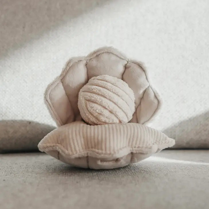The LAMBWOLF COLLECTIVE - OYSTER POP DOG TOY by LAMBWOLF COLLECTIVE is a soft, plush toy representing a light beige shell with a round, textured pearl inside. Crafted from a corduroy-like material, the shell and pearl showcase multiple textures, giving them a cozy and inviting appearance. This Sea World Edition toy is displayed against a neutral fabric background.