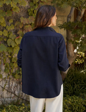 A woman stands barefoot indoors, smiling with her hands in her pockets, dressed in the FRANK & EILEEN EILEEN Relaxed Button Up Shirt in Italian Cashmere Touch Navy and cream wide-leg pants. Sunlight streams through sheer curtains, casting soft shadows across the floor.