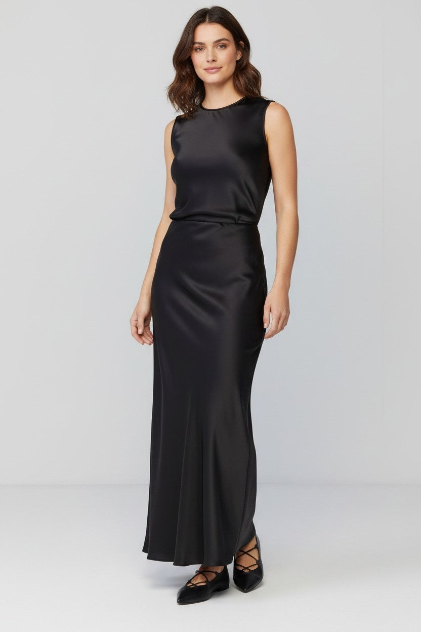 A woman stands smiling, wearing the MILVA MI - LONG SATIN SKIRT IN BLACK by MILVA, styled with a fluid silhouette. The skirt flows to the floor in lustrous black satin, paired with a fitted top that features long sleeves and a cowl neckline. She has wavy brown hair and wears black shoes, standing against a plain light background.