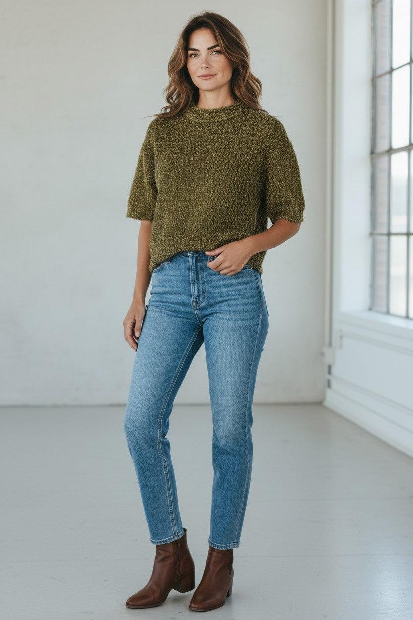 A woman stands indoors wearing the FLOOR - SHORT SLEEVE SHIMMER SWEATER IN RESIN by FLOOR, a textured olive green short-sleeve sweater crafted from a soft viscose-blend knit. She pairs it with light blue jeans and brown ankle boots. Her hair falls in loose waves and she has a neutral expression, set against a minimal background of white walls and a window.