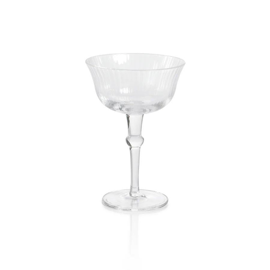 The JULIEN CLEAR OPTIC MARTINI GLASS by ZODAX sits on a reflective white surface, showcasing its transparent glass construction. The glass features a short stem and a large, wide bowl with a 120 ml capacity. Subtle vertical ribbing on the bowl lends it a classic, elegant appearance.