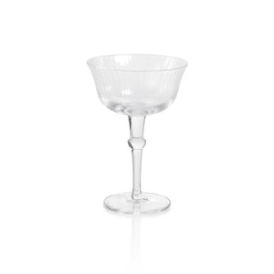 The JULIEN CLEAR OPTIC MARTINI GLASS by ZODAX sits on a reflective white surface, showcasing its transparent glass construction. The glass features a short stem and a large, wide bowl with a 120 ml capacity. Subtle vertical ribbing on the bowl lends it a classic, elegant appearance.