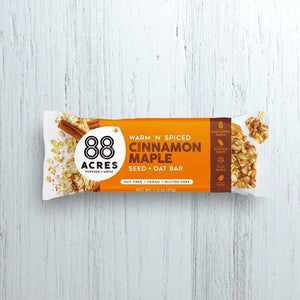 Image of an 88 ACRES Cinnamon Maple Seed + Oat Bar. The packaging is white with an orange section that displays the brand "88 ACRES" and the flavor details. The package showcases the nut-free, vegan, and gluten-free seed bar sprinkled with oats and seeds.
