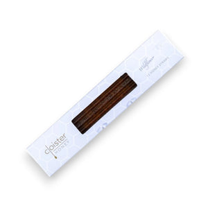 A floral teacup filled with tea sits on a square blue plate with two rectangular biscuits and an open Cloister Honey - Wildflower Honey Straw. A box labeled "Artisan Honey" and a jar containing more Wildflower Honey Straws are beside the plate on a rustic wooden surface with a grassy background.