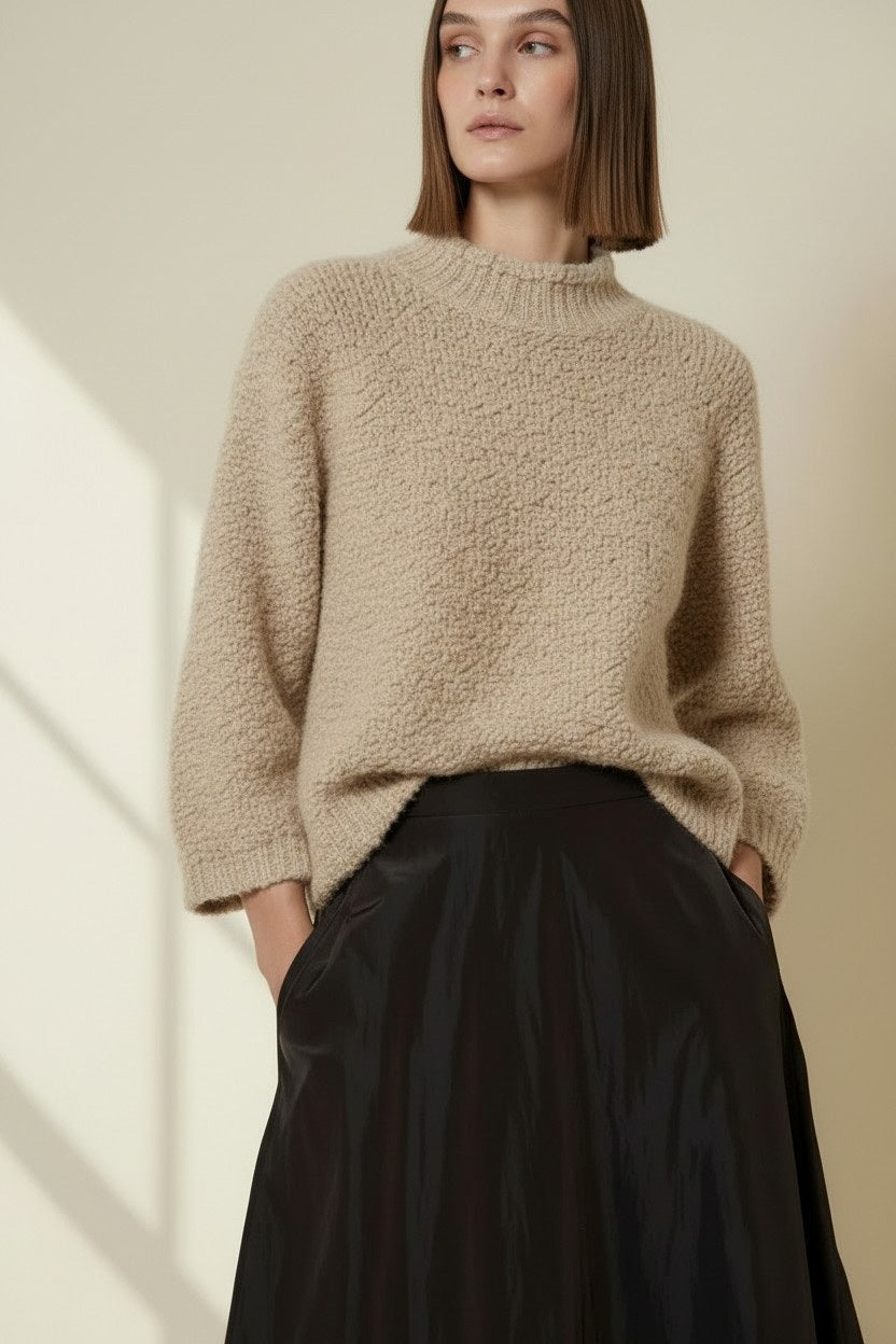 A woman stands indoors by a window, wearing the VICARIO CINQUE - MADEIRA Boucle Sweater in Light Taupe from VICARIO CINQUE, paired with a long black skirt and sneakers. Sunlight casts grid-like shadows on the wall.