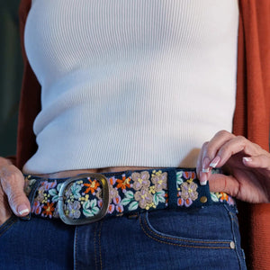JENNY KRAUSS - MORNING DEW FLORAL EMBROIDERED WOOL BELT in dark blue wool with colorful floral embroidery and a silver-tone metal buckle, worn over blue jeans; this handcrafted Peruvian accessory highlights intricate needlework.