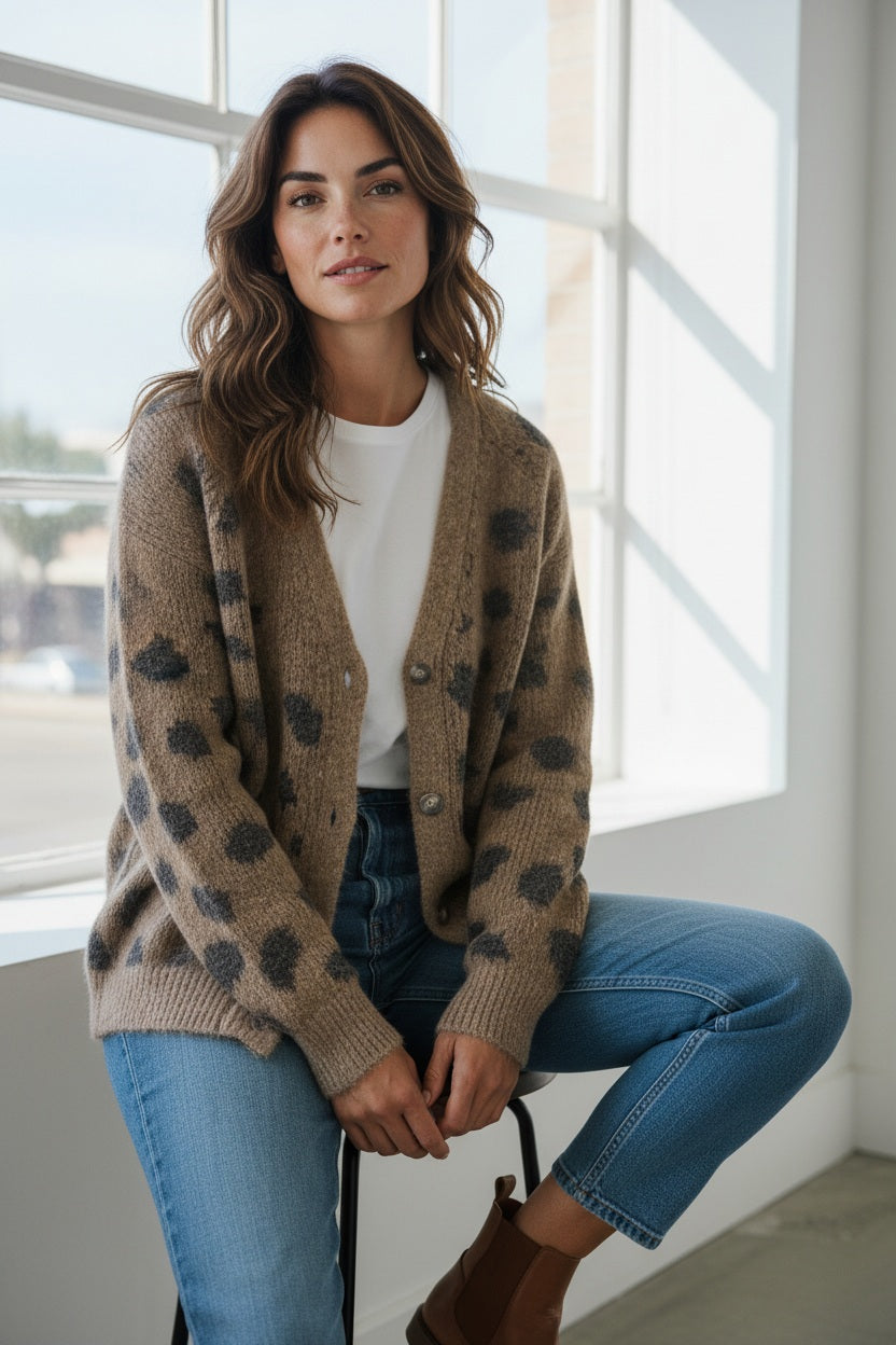 A woman with wavy brown hair sits on a stool by a large window, wearing the HARTFORD - MONSON WOOL CARDIGAN by Hartford—a casual chic brown women’s cardigan with black spots—layered over a white t-shirt, paired with blue jeans and brown ankle boots. She looks relaxed and faces the camera.