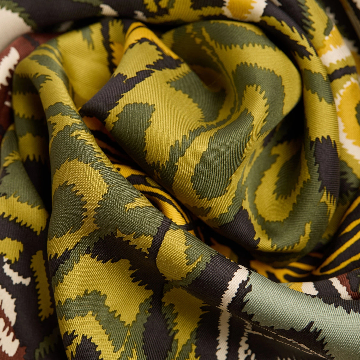 A close-up of a woman wearing a beige blazer and the INOUI EDITIONS - PAPILLON (BUTTERFLIES) Silk Scarf in Green, featuring abstract patterns in yellow, black, green, and white. Only her lower face and neck are visible.