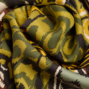 A close-up of a woman wearing a beige blazer and the INOUI EDITIONS - PAPILLON (BUTTERFLIES) Silk Scarf in Green, featuring abstract patterns in yellow, black, green, and white. Only her lower face and neck are visible.