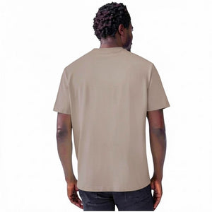 A man with short curly hair and a beard wears the MACK WELDON Heavyweight Slub Cotton Pocket T-Shirt in Desert Taupe and dark pants, standing against a plain white background.