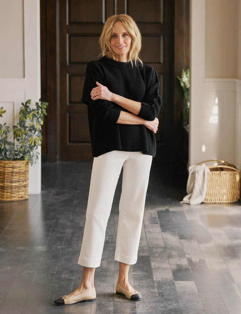 Wearing FRANK & EILEEN’s WESTPORT ILLUSION PULL ON WIDE LEG JEAN in Antique White, a person pairs them with a black sweater and beige flats with black toes, standing on a dark wooden floor in a bright, modern room.