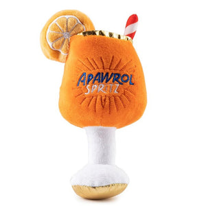 A fluffy Pomeranian dog sits on a rug outdoors next to the HAUTE DIGGITY DOG APAWROL SPRITZ DOG TOY, a squeaky toy shaped like an orange cocktail with a straw and lemon slice.